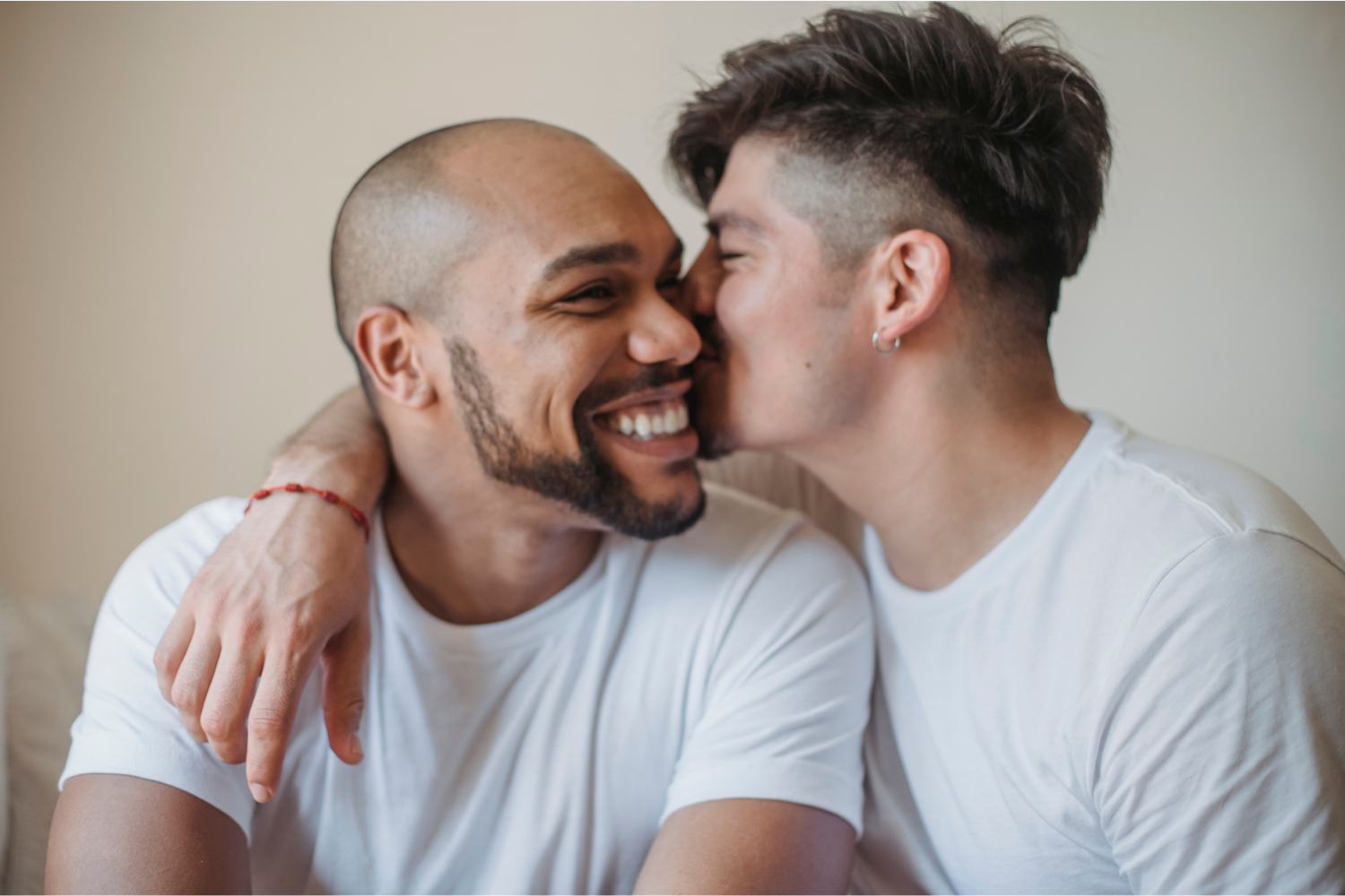 Two men in white t-shirts, one man is kissing the other man's cheek whilst he smiles