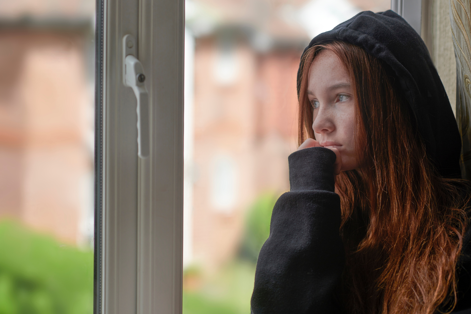 Teenager in a black hoodie looking out of the window with a sad look on her face