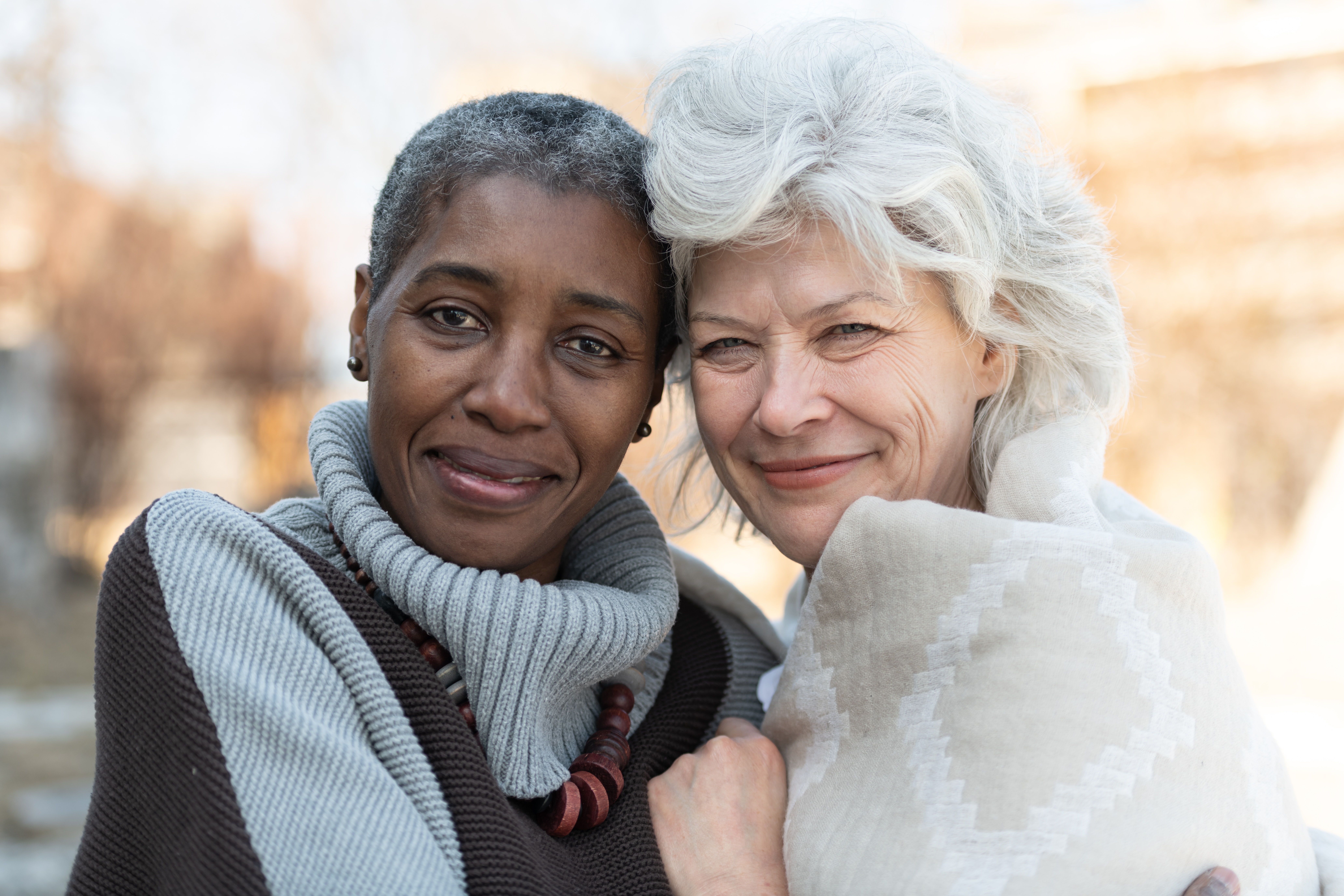 Two senior friends laugh affectionately together. The women are standing outside on a sunny but cool day. They are dressed in casual sweaters. One woman is of African descent and the other is Caucasian.