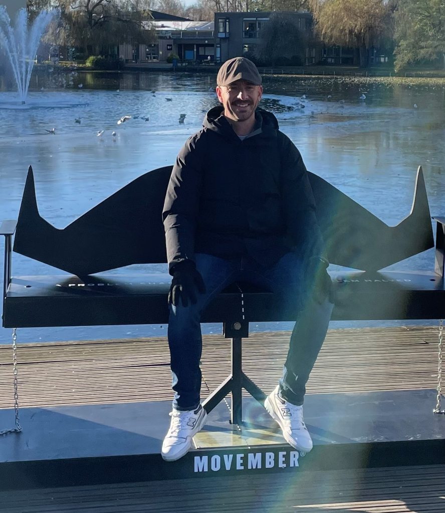 A man in casual clothing, Professor Paul Galdas, sat on top of a giant life-sized moustache with 'Movember' written underneath it. He is smiling and there is water